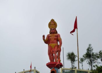 Statue of Lord Hanuman standing with a raised hand and mace, adorned with gold ornaments and red flags, at a Hindu temple