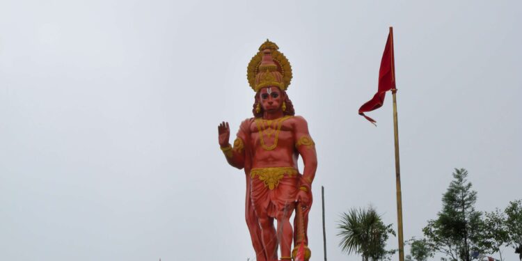 Statue of Lord Hanuman standing with a raised hand and mace, adorned with gold ornaments and red flags, at a Hindu temple