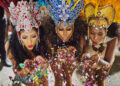 Three women in elaborate red, blue, and gold carnival headdresses blowing colourful confetti towards the camera.