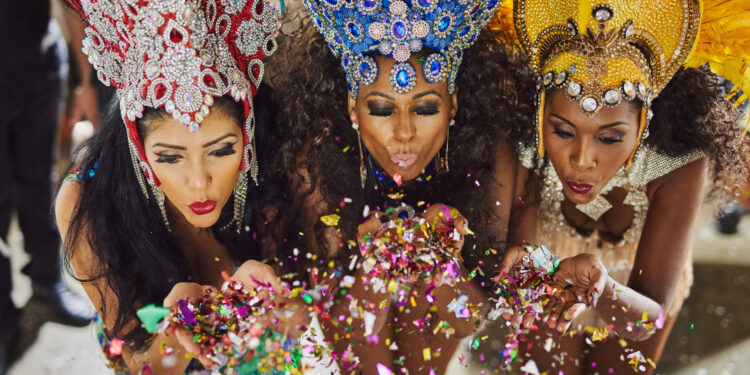 Three women in elaborate red, blue, and gold carnival headdresses blowing colourful confetti towards the camera.