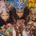 Three women in elaborate red, blue, and gold carnival headdresses blowing colourful confetti towards the camera.
