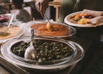 A person serving Caribbean-style stewed greens, lentils, and vegetables onto a plate with roti.