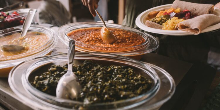 A person serving Caribbean-style stewed greens, lentils, and vegetables onto a plate with roti.
