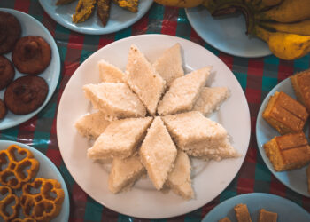 Assorted Indian mithai and Caribbean desserts arranged on plates, including coconut barfi, rum cake, and fried sweets.