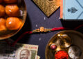 Traditional Raksha Bandhan items arranged on a dark background, including a red rakhi, Indian sweets (motichoor laddoos), a 500 rupee note, a golden thali with diya, rice, kumkum, and roli.