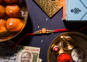 Traditional Raksha Bandhan items arranged on a dark background, including a red rakhi, Indian sweets (motichoor laddoos), a 500 rupee note, a golden thali with diya, rice, kumkum, and roli.