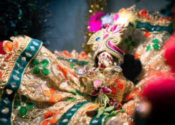 Close-up of a decorative idol of Hindu god Lord Krishna, adorned in vibrant, intricate clothing with gold embroidery, colourful gemstones, and a peacock-feather-style headpiece.