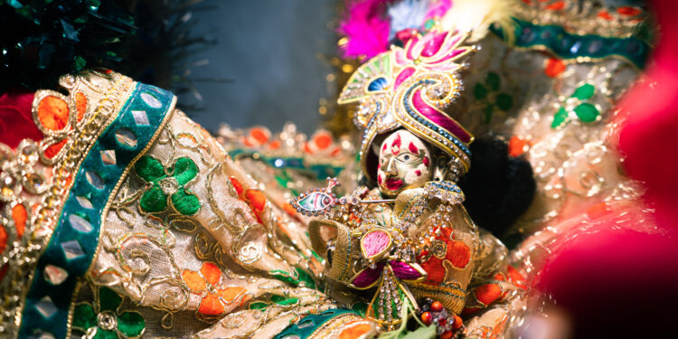 Close-up of a decorative idol of Hindu god Lord Krishna, adorned in vibrant, intricate clothing with gold embroidery, colourful gemstones, and a peacock-feather-style headpiece.