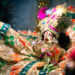 Close-up of a decorative idol of Hindu god Lord Krishna, adorned in vibrant, intricate clothing with gold embroidery, colourful gemstones, and a peacock-feather-style headpiece.