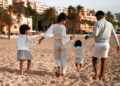 Indo-Caribbean family walking together on a sandy beach, holding hands and dressed in coordinated light-coloured outfits.