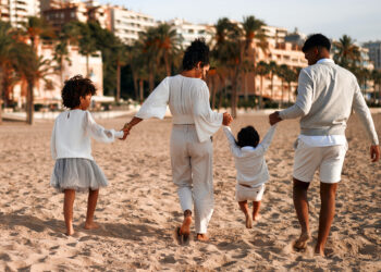 Indo-Caribbean family walking together on a sandy beach, holding hands and dressed in coordinated light-coloured outfits.