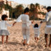 Indo-Caribbean family walking together on a sandy beach, holding hands and dressed in coordinated light-coloured outfits.
