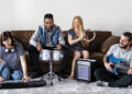 Musicians jamming with keyboard, drum, tambourine, and electric guitar in a cosy living room.