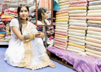 Woman in white and gold sari sitting in a colourful fabric shop, wearing traditional Indo-Caribbean jewellery.