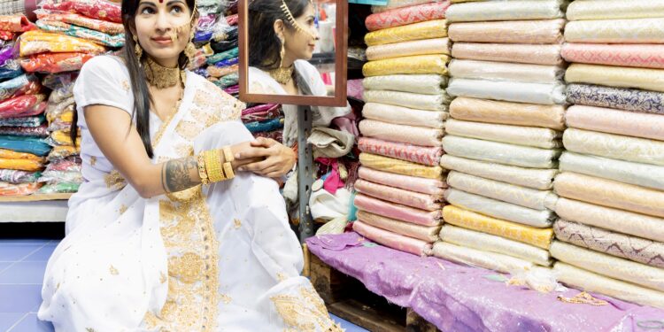 Woman in white and gold sari sitting in a colourful fabric shop, wearing traditional Indo-Caribbean jewellery.