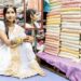 Woman in white and gold sari sitting in a colourful fabric shop, wearing traditional Indo-Caribbean jewellery.
