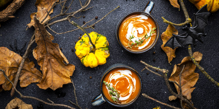 Two mugs of creamy pumpkin soup with thyme garnish surrounded by autumn leaves, gourds, and rustic branches — perfect for Indo-Caribbean autumn warmers.