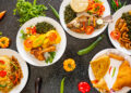 Plates of vibrant Afro-Caribbean and Indo-Caribbean dishes with rice, fried fish, chicken, curries, roti, and fresh vegetables, arranged on a dark table.