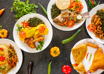 Plates of vibrant Afro-Caribbean and Indo-Caribbean dishes with rice, fried fish, chicken, curries, roti, and fresh vegetables, arranged on a dark table.