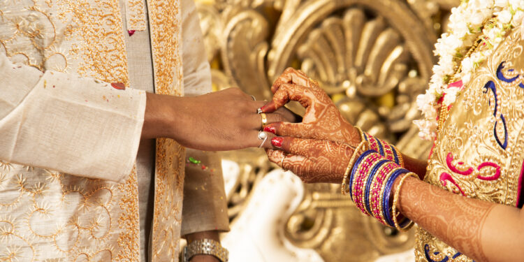 A vibrant Indo-Caribbean wedding in the UK, featuring a couple beneath a mandap decorated with marigolds and tropical flowers, surrounded by guests in colourful attire blending Indian and Caribbean styles.