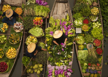 Aerial view of colourful floating market boats filled with fresh fruits, vegetables, herbs, and flowers, with vendors wearing traditional straw hats on a canal.