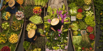 Aerial view of colourful floating market boats filled with fresh fruits, vegetables, herbs, and flowers, with vendors wearing traditional straw hats on a canal.