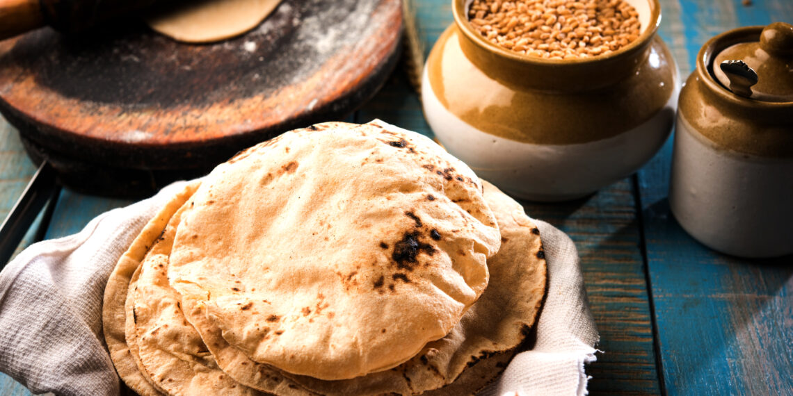 Stack of freshly cooked Caribbean rotis with light char marks resting in a basket beside a rolling board, wheat grains, and a clay jar on a rustic wooden table.