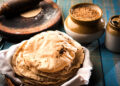 Stack of freshly cooked Caribbean rotis with light char marks resting in a basket beside a rolling board, wheat grains, and a clay jar on a rustic wooden table.