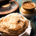 Stack of freshly cooked Caribbean rotis with light char marks resting in a basket beside a rolling board, wheat grains, and a clay jar on a rustic wooden table.