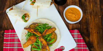 Caribbean-style roti wrap filled with spiced chicken, green beans and grilled aubergine on a white board with dipping sauce