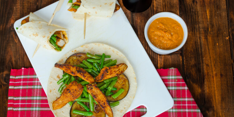 Caribbean-style roti wrap filled with spiced chicken, green beans and grilled aubergine on a white board with dipping sauce
