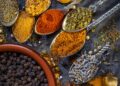 Assorted Indo-Caribbean spice blends and whole spices displayed in metal spoons, including turmeric, chilli powder, dried herbs, and black peppercorns on a rustic surface.