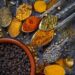 Assorted Indo-Caribbean spice blends and whole spices displayed in metal spoons, including turmeric, chilli powder, dried herbs, and black peppercorns on a rustic surface.