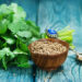 Fresh cilantro leaves beside a wooden bowl filled with coriander seeds on a rustic blue wooden surface.