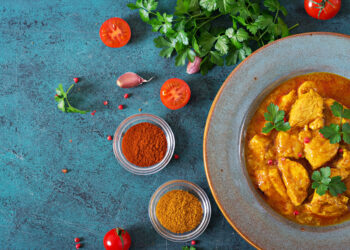 Bowl of golden chicken curry garnished with fresh parsley, surrounded by spices, tomatoes, garlic, and herbs on a blue textured surface.