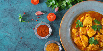 Bowl of golden chicken curry garnished with fresh parsley, surrounded by spices, tomatoes, garlic, and herbs on a blue textured surface.