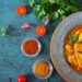 Bowl of golden chicken curry garnished with fresh parsley, surrounded by spices, tomatoes, garlic, and herbs on a blue textured surface.
