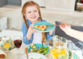 Smiling young girl with braided hair holding a plate with a slice of pie while sitting at a family dinner table with salad, juice, and infused water.