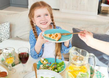 Smiling young girl with braided hair holding a plate with a slice of pie while sitting at a family dinner table with salad, juice, and infused water.
