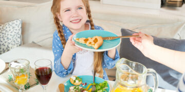 Smiling young girl with braided hair holding a plate with a slice of pie while sitting at a family dinner table with salad, juice, and infused water.
