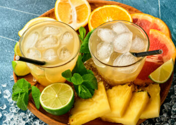 Two glasses of chilled citrus tropical drinks with ice cubes, surrounded by sliced pineapple, lime, orange, grapefruit, and fresh mint on a wooden tray.