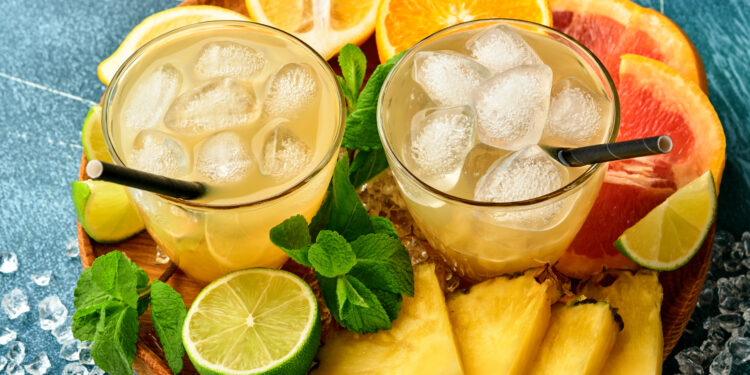 Two glasses of chilled citrus tropical drinks with ice cubes, surrounded by sliced pineapple, lime, orange, grapefruit, and fresh mint on a wooden tray.