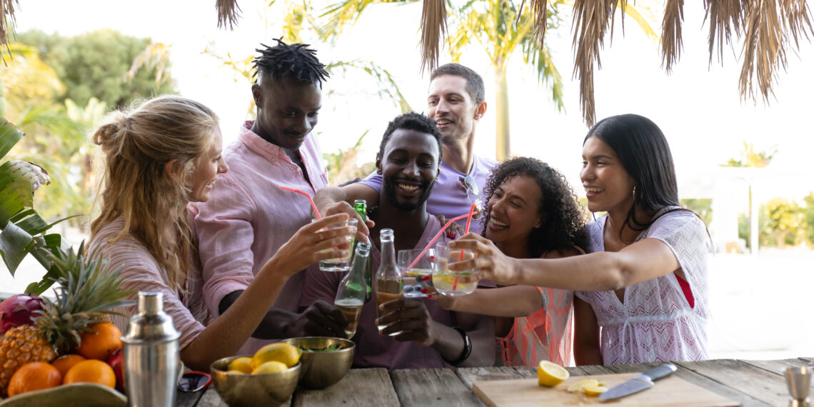 Group of friends toasting drinks at a tropical outdoor table while preparing fresh cocktails and fruit.