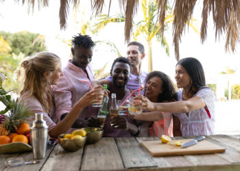 Group of friends toasting drinks at a tropical outdoor table while preparing fresh cocktails and fruit.