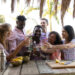 Group of friends toasting drinks at a tropical outdoor table while preparing fresh cocktails and fruit.