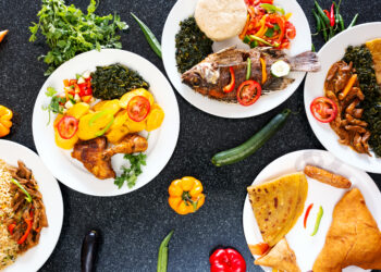Assorted traditional Caribbean-style dishes served on white plates, including curry chicken with potatoes, fried fish, stewed meat, rice, roti, and sautéed greens, arranged on a dark tabletop with fresh vegetables and herbs.