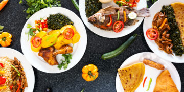 Assorted traditional Caribbean-style dishes served on white plates, including curry chicken with potatoes, fried fish, stewed meat, rice, roti, and sautéed greens, arranged on a dark tabletop with fresh vegetables and herbs.