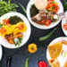 Assorted traditional Caribbean-style dishes served on white plates, including curry chicken with potatoes, fried fish, stewed meat, rice, roti, and sautéed greens, arranged on a dark tabletop with fresh vegetables and herbs.