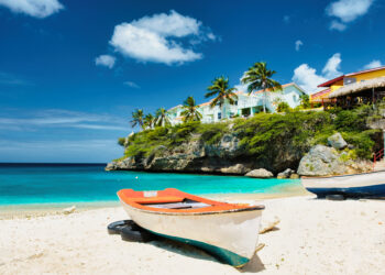 Colourful fishing boats resting on a quiet Caribbean beach with turquoise water, palm trees, and cliffside houses under a bright blue sky, reflecting a peaceful heritage travel atmosphere.