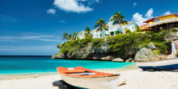 Colourful fishing boats resting on a quiet Caribbean beach with turquoise water, palm trees, and cliffside houses under a bright blue sky, reflecting a peaceful heritage travel atmosphere.